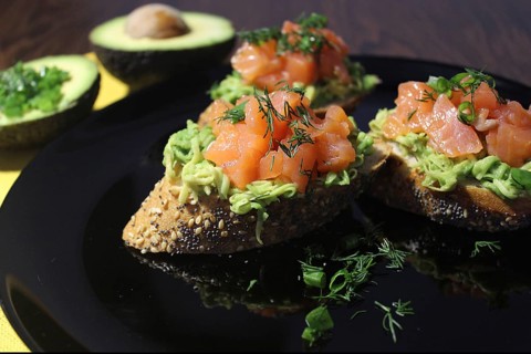 Three slices of seeded bread topped with avocado, salmon, and dill on a black plate with avocado