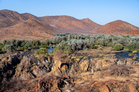 Kunene River with green vegetation in dry red mountain landscape, waterfall and African Baobab
