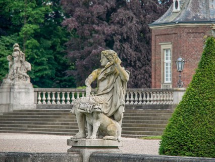 Stone sculpture in front of a castle with stone stairs and balustrades, surrounded by green bushes