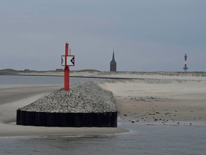 Red beacon on gravel embankment, divided coastal landscape with sand dunes and church tower in the