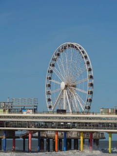 Ferris wheel on a pier reaching into the sea under a blue sky, parasols and a pier on the beach of