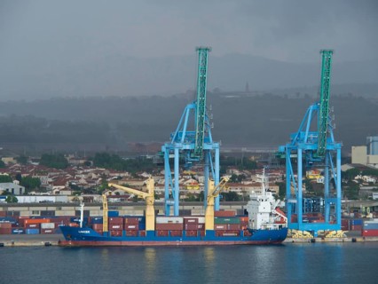 A freighter stands in the harbour with large blue cranes and many containers in the background,