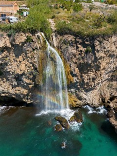 A waterfall cascades from a cliff into turquoise-coloured water in a rocky, natural setting, aerial