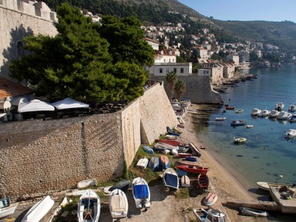 Boats moored on the shore next to a city wall, the sun shining brightly and the clear sky adding to