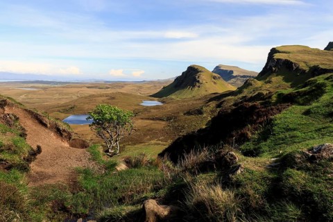 A wide view of a hilly landscape with a path, small lakes and green grass under a clear sky,