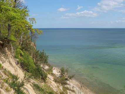 A steep rocky outcrop with green trees, a path leading down to the calm sea under a clear sky,