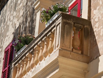 A small balcony with flowers and shutters adorns an old stone wall, the town of mdina on the island