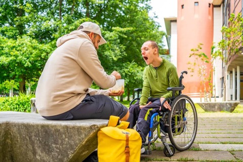 Male caucasian adult university student feeding a friend with cerebral palsy sitting on wheelchair