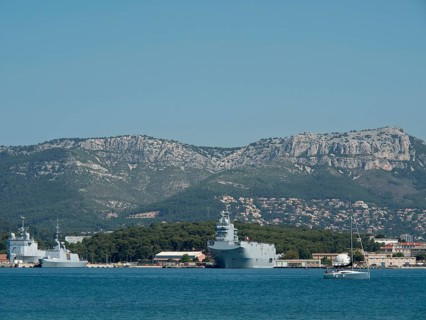 Several warships and a sailing boat in the harbour, green coast and mountain range in the