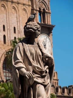 Stone statue with a dove on its head, a clock tower rises in the background, palermo in sicily with