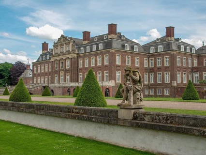 Historic castle with manicured lawn, statues and bushes under a cloudy sky and spacious entrance,