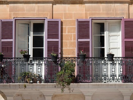 A mediterranean balcony with purple shutters and potted plants in front of two open windows, the