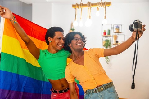 Happy gay couple celebrating diversity taking selfie at home raising LGBT rainbow flag