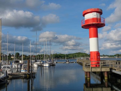 Tall red and white lighthouse at the edge of a harbour with calm water, surrounded by sailing