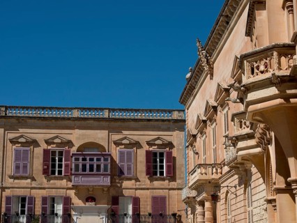 Two buildings with historic stone walls and shutters under a clear sky, the town of mdina on the