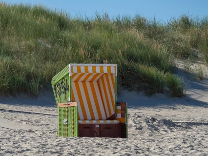 A single green beach chair with yellow and white stripes stands on the beach in front of a grassy