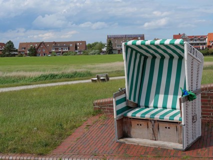 A green and white beach chair stands on a paved square with houses in the background, Baltrum