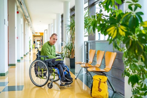 Man with cerebral palsy in wheelchair in the university waiting in the corridor next to school bag