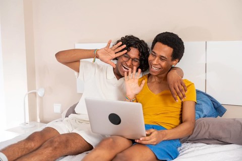 Gay couple waving during a video call with laptop lying on bed