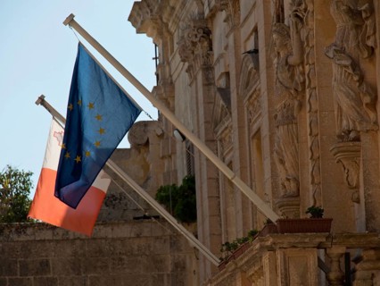 Two flags, one of the EU and one of Malta, in front of historic stone architecture, the town of