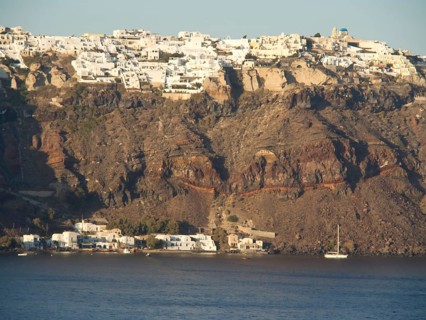A cluster of white buildings on a cliff dramatically jutting out over the calm sea, brown volcanic