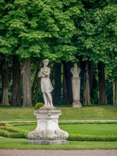 A statue in a green park with tall trees and another monument in the background, historic castle