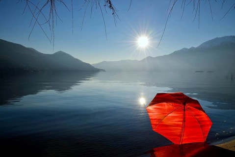 Red Umbrella Leaning on the Sand Beach on Alpine Lake Maggiore with Mountain and Sunbeam and