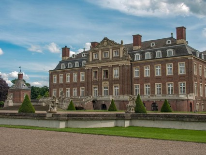 Historic castle with manicured lawn and sculptures, surrounded by a stone wall under a cloudy sky,