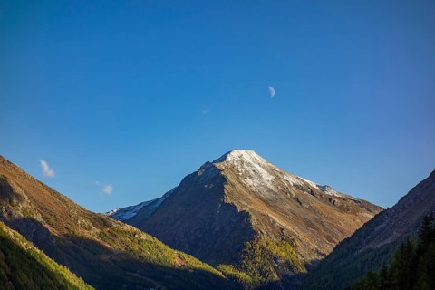 A snow-covered mountain peak under a blue sky with a small crescent moon, surrounded by mountains