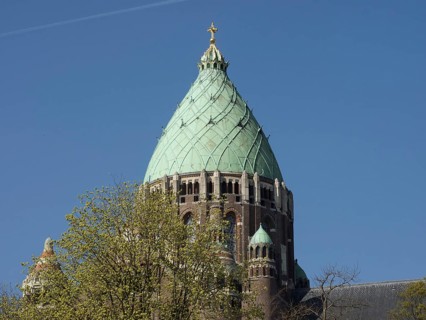 The upper part of a church tower with a greenish dome and a golden cross surrounded by tree tops,