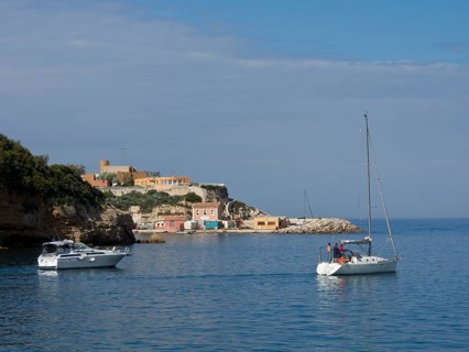 Sailing boats in front of a coastal landscape with cliffs and small houses under a partly cloudy