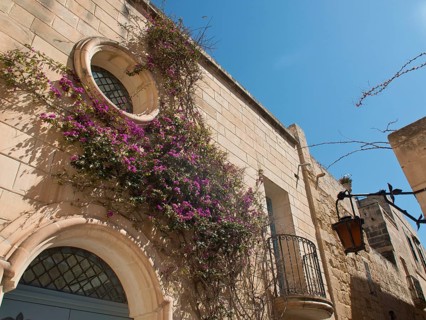 Part of a stone building with round windows and a small balcony, surrounded by purple flowers and