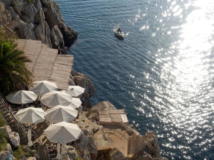 View of the sea from the rock, with white parasols and a boat on the water, reflecting the sun's