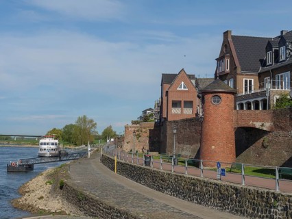 Historic brick buildings along a riverside footpath in sunny weather with a slightly cloudy sky,