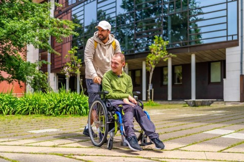 University student pushing the wheelchair of a disabled friend outdoors in the university campus