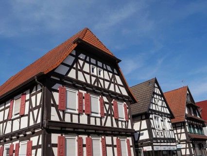 Three half-timbered houses with red shutters and tiled roofs, under a blue sky in the sun, historic
