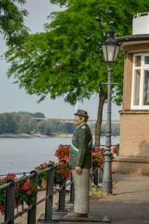 Statue of a man in uniform next to a walkway on the riverbank with lamps and blooming flowers,