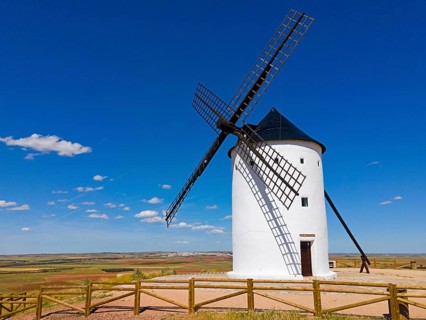 A windmill stands alone in a field under a bright blue sky in a vast summer landscape, aerial view,