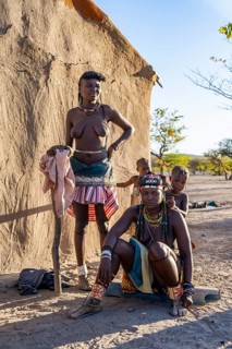 Two Hakaona woman with traditional kapapo hairstyle, with children, in front of their mud hut, in