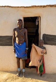 Hakaona woman with traditional kapapo hairstyle and colourful necklaces, with child, in front of