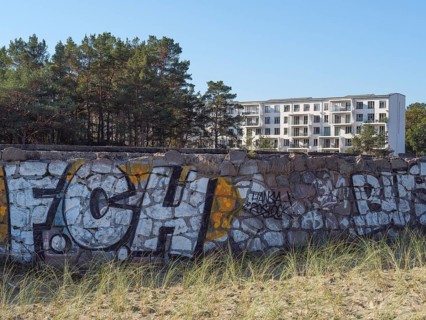 A stone wall with colourful graffiti in front of a modern building, framed by pine trees and tufts