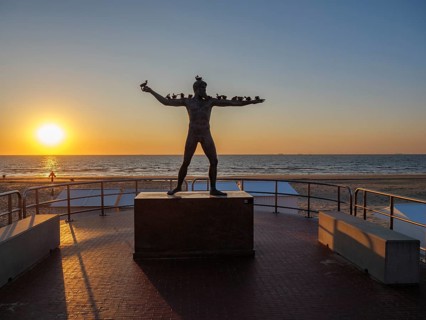 A statue in front of a sunset on the beach, surrounded by a railing, sunset in many colours on the