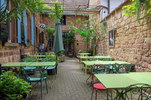 A cosy outdoor area of a cafe with green tables and chairs, surrounded by plants and brick walls,