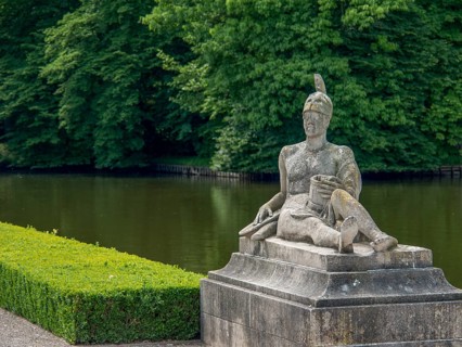 Stone sculpture of a seated man next to a pond, surrounded by lush greenery and trees, old red