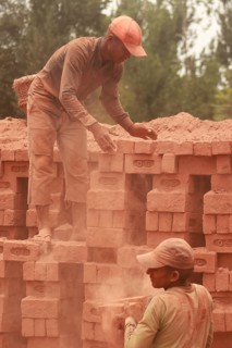 Two workers stacking red bricks in a dusty outdoor setting, both wearing hats and working together,