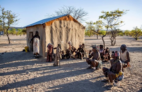 Traditional Hakaone woman and men sit in front of a mud house in their village, in the morning