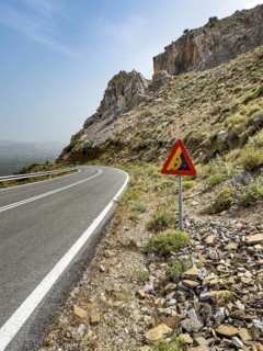 Warning sign for falling rocks next to mountain road near Plora in the background rocky mountains,