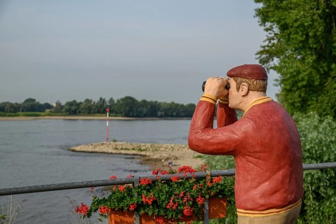 Man statue with binoculars on the riverside, flowers in the foreground and trees in the background,