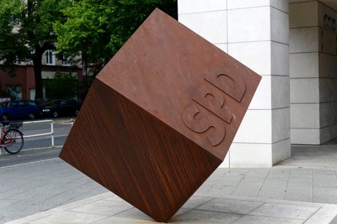 Red-brown cube with SPD inscription in front of a modern building on a street with a bicycle in the