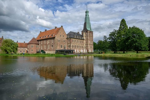 Large castle with brick walls and towers by a pond with reflections, surrounded by trees under a